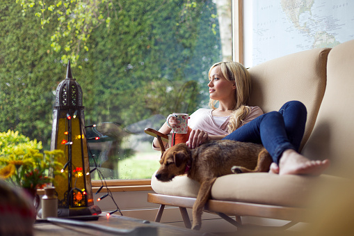 A happy blonde woman lies down on her sofa at home with a hot drink in her hand while looking out of her window. Her dog is lying next to her asleep. Ornaments and lights can be seen in the room.